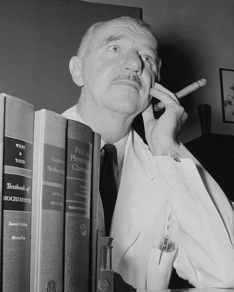 Vincent du Vigneaud seated beside a bookcase, holding a cigar
